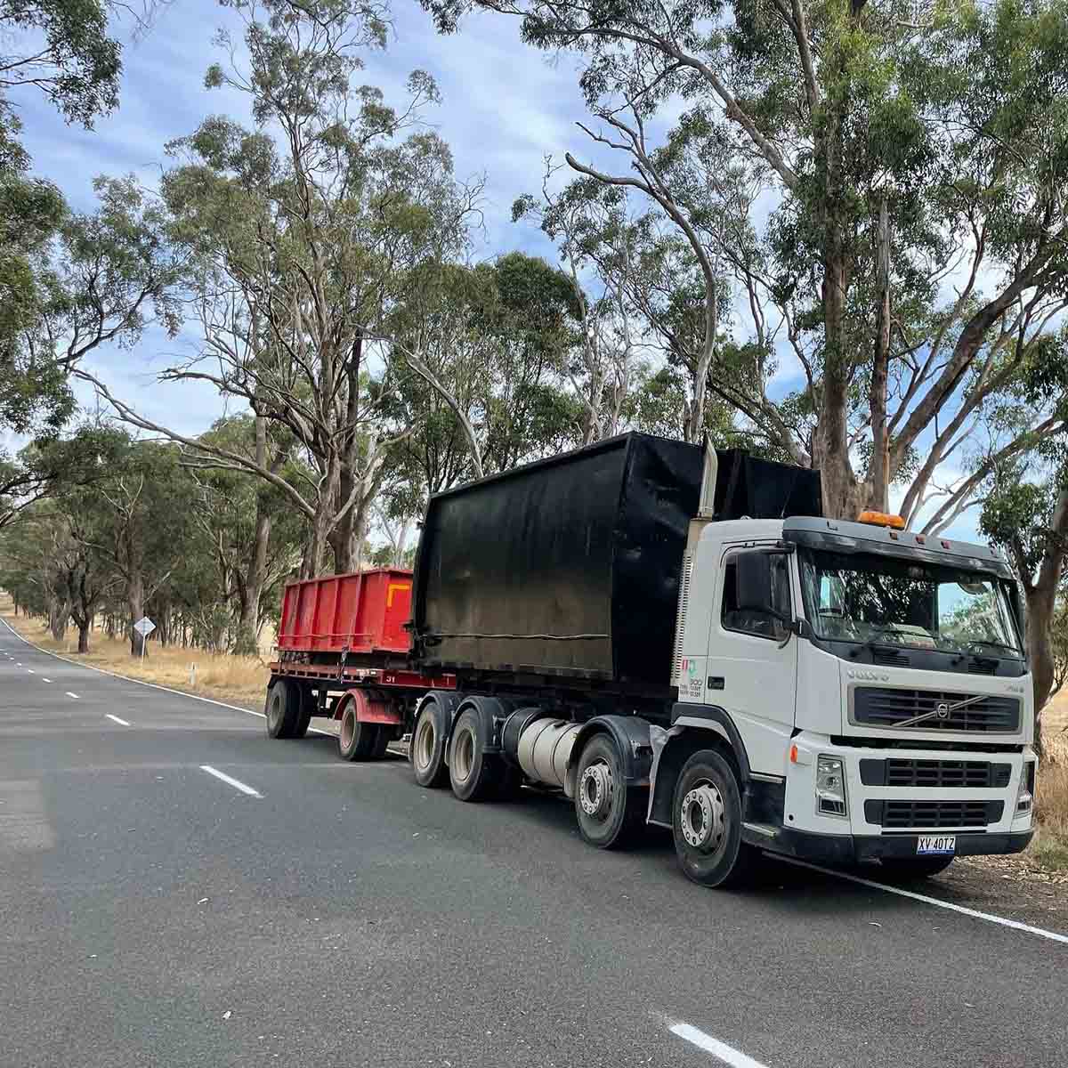 White truck carrying a black bin and towing a red bin on a trailer along a tree-lined road