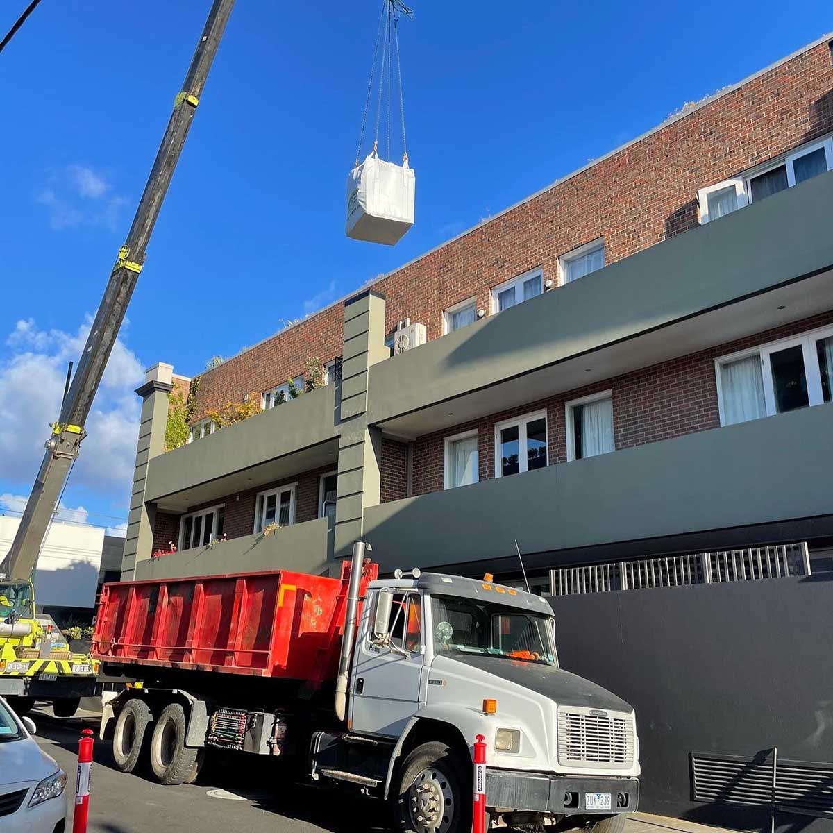 Truck with a red bin parked outside an apartment building while a crane lifts a load