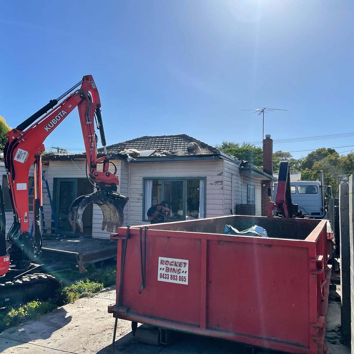 Mini excavator with a grab demolishing a weatherboard house beside a red bin