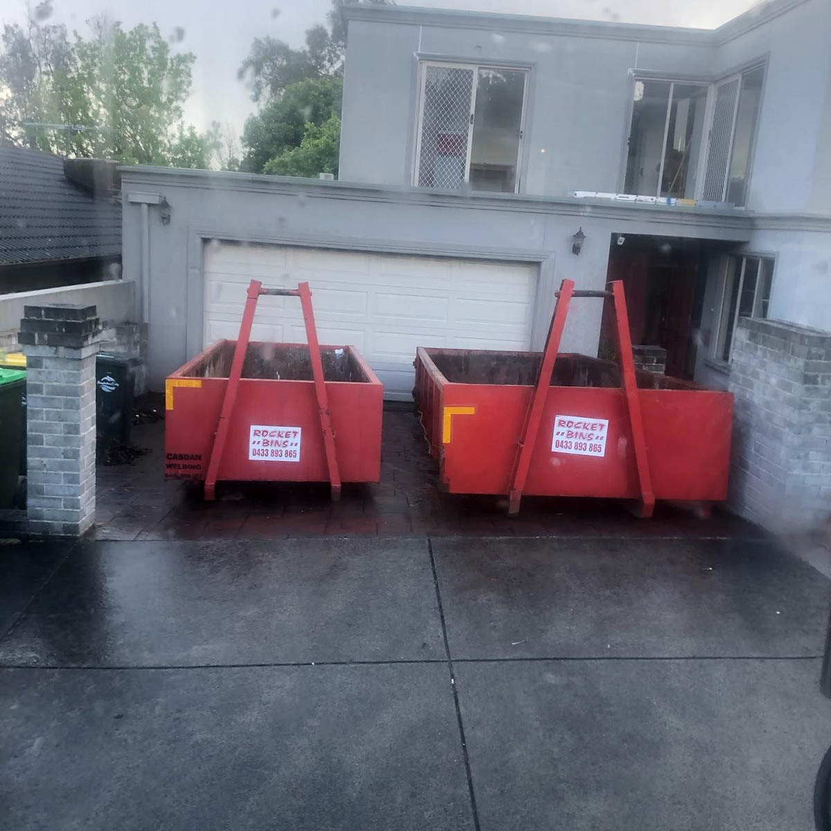 Two red walk-in bins on a concrete driveway in front of a house